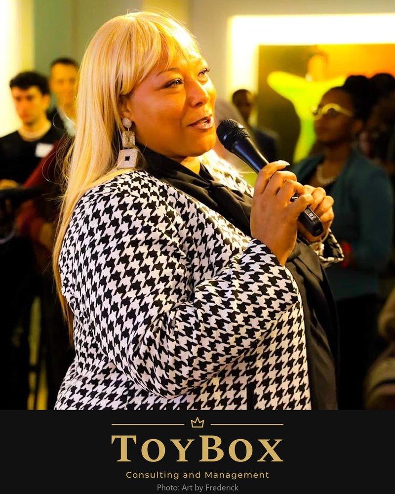 Katoya Palmer speaking with a microphone, wearing a houndstooth jacket, above the ToyBox Consulting and Management logo in gold