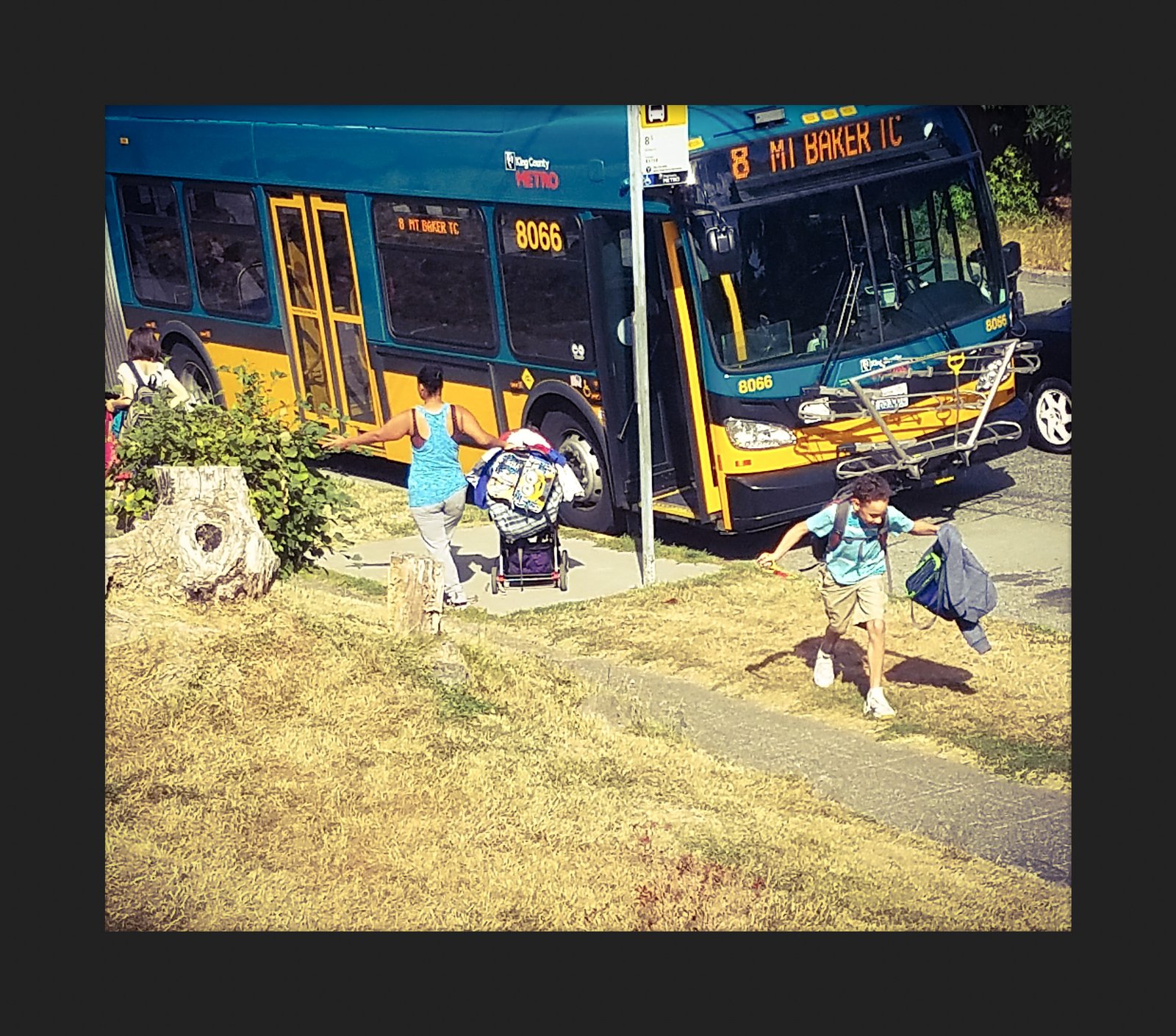 A city bus at a stop beside a grassy lot, with people walking past