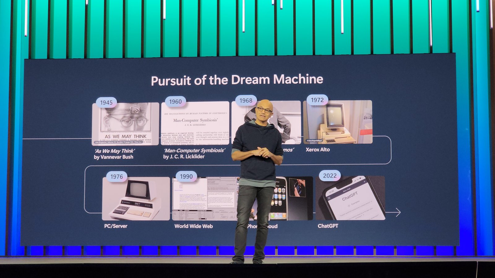 A speaker on a stage in front of a screen titled Pursuit of the Dream Machine