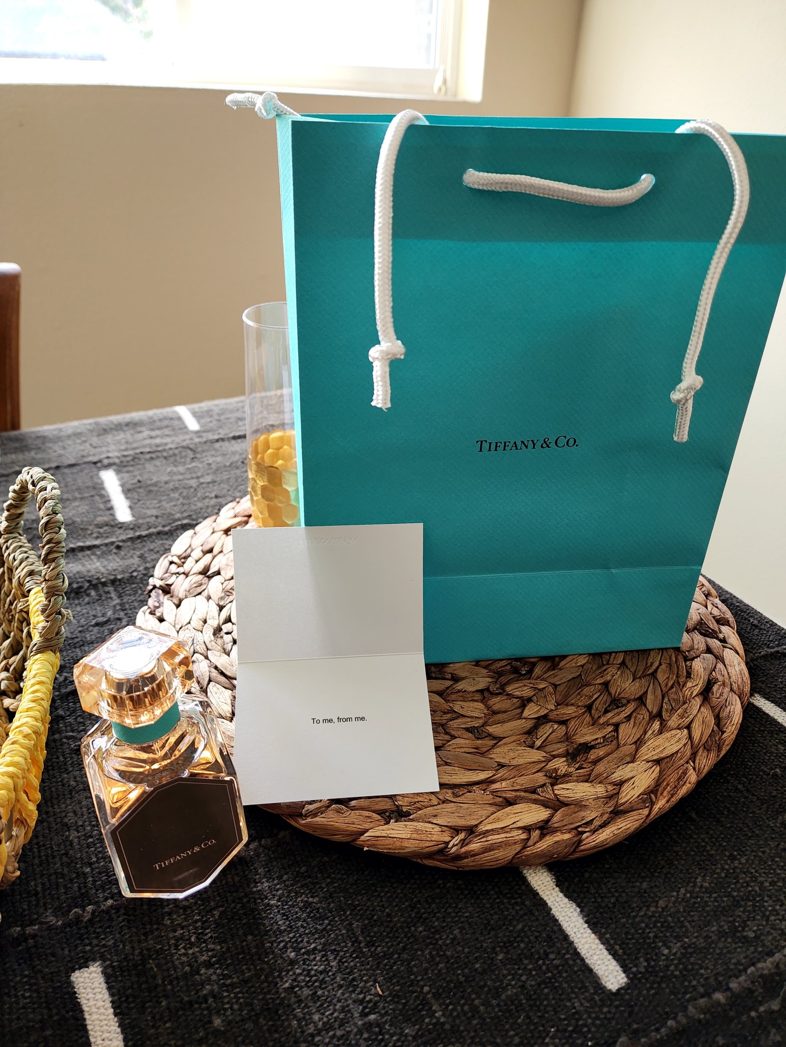 A turquoise gift bag, a small card and a perfume bottle on a woven tray