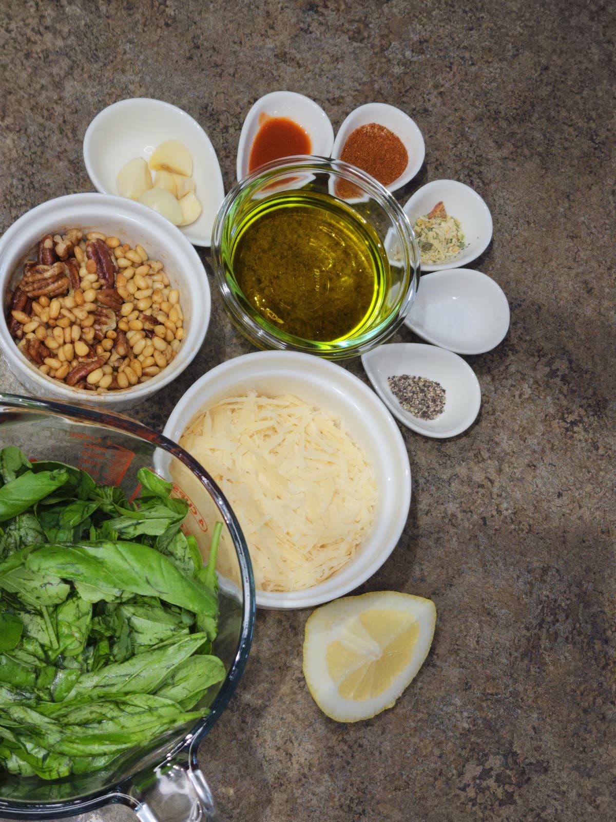 Cajun pesto ingredients laid out: fresh basil, olive oil, pecans and pine nuts, parmesan, garlic, half a lemon and small bowls of spices