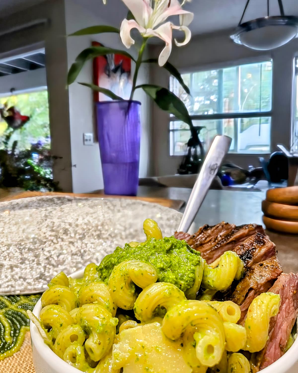 A bowl of cavatappi tossed in Cajun pesto with sliced grilled steak, a purple vase and lily behind it